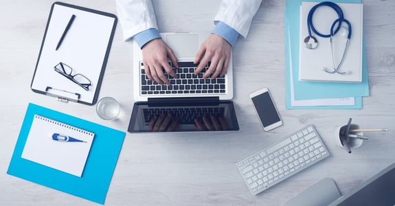 Arztschreibtisch mit Laptop und weiteren Arbeitsmaterialien, symbolisiert die Digitalisierung im medizinischen Bereich.